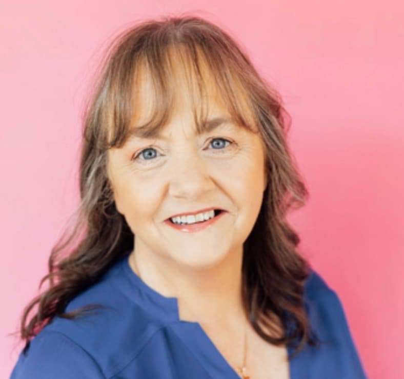 Smiling woman with brown hair and blue eyes wearing blue against a bright pink background.