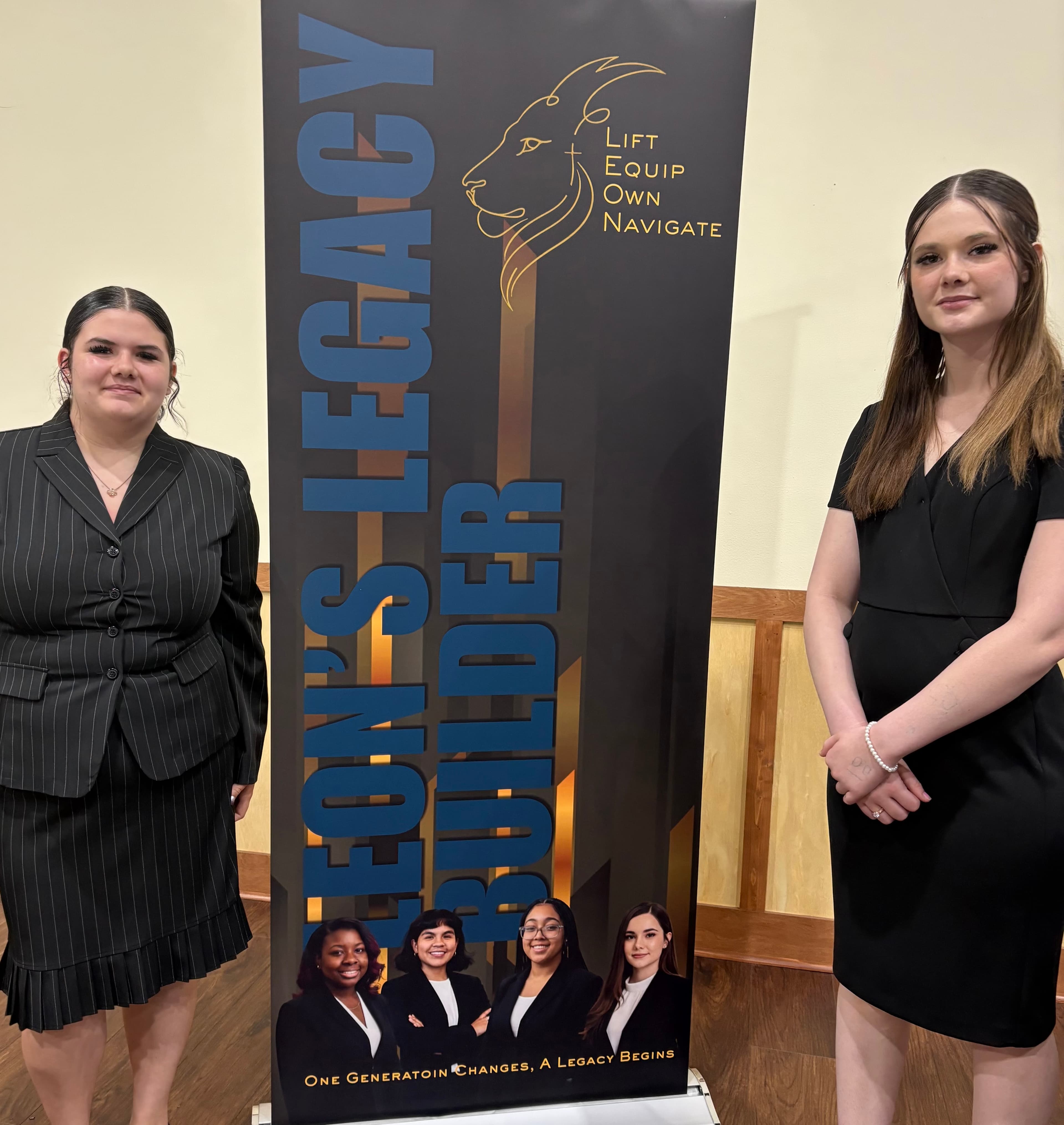 Two young women in professional black attire stand beside a Leon's Legacy Builder banner.