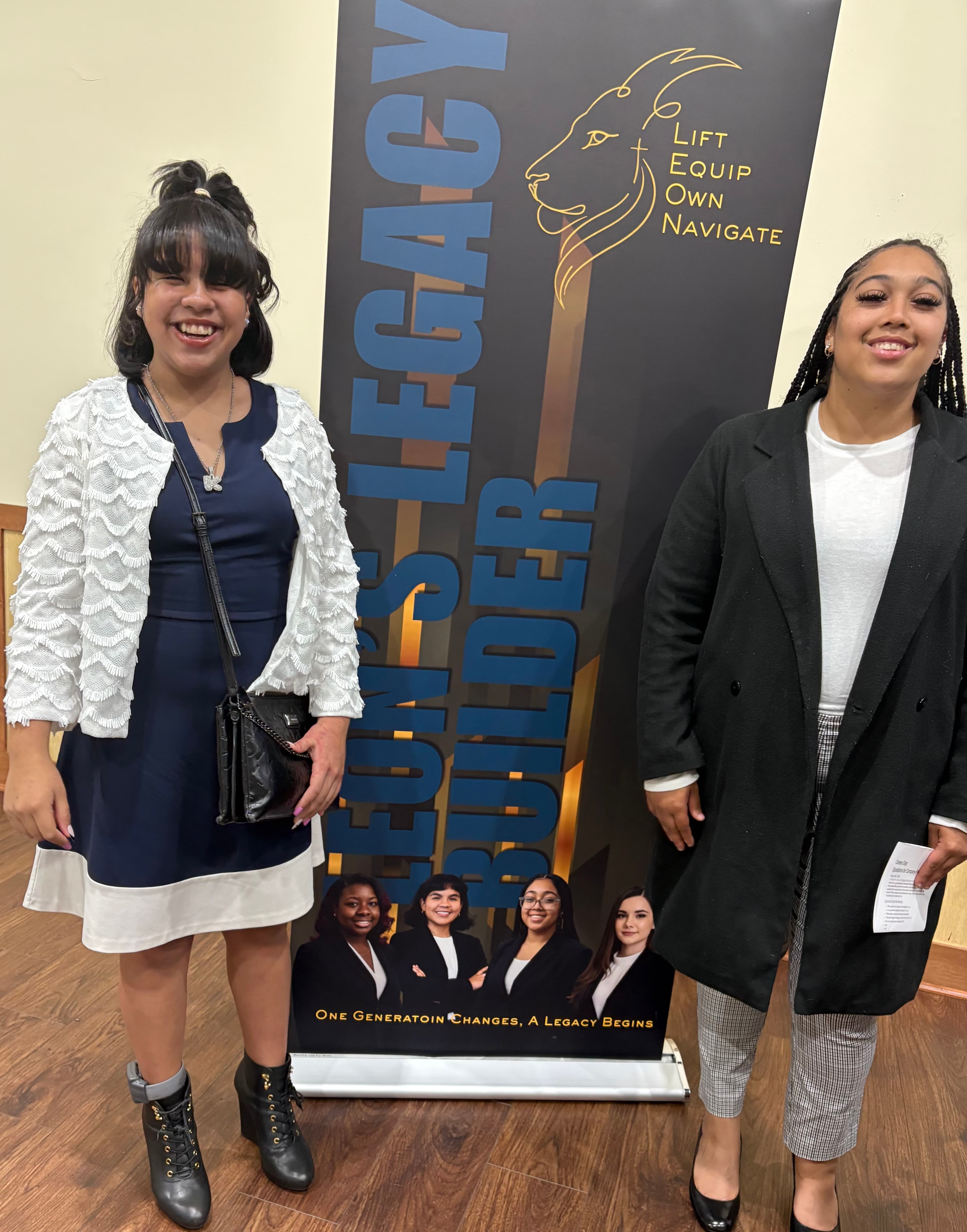 Two smiling young women pose beside a banner for the Leon's Legacy Builder program.