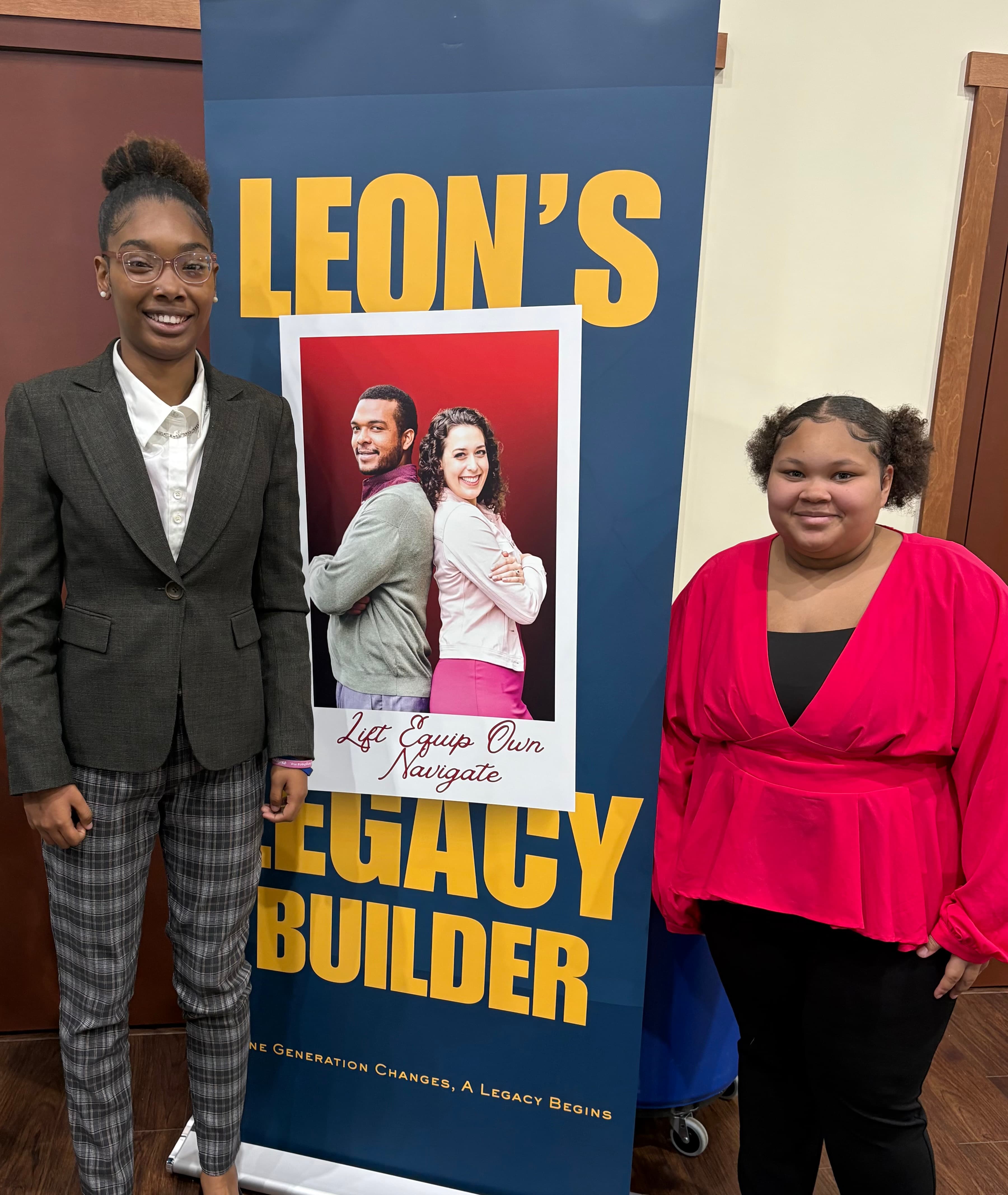 Two young women pose on either side of a blue Leon's Legacy Builder banner.
