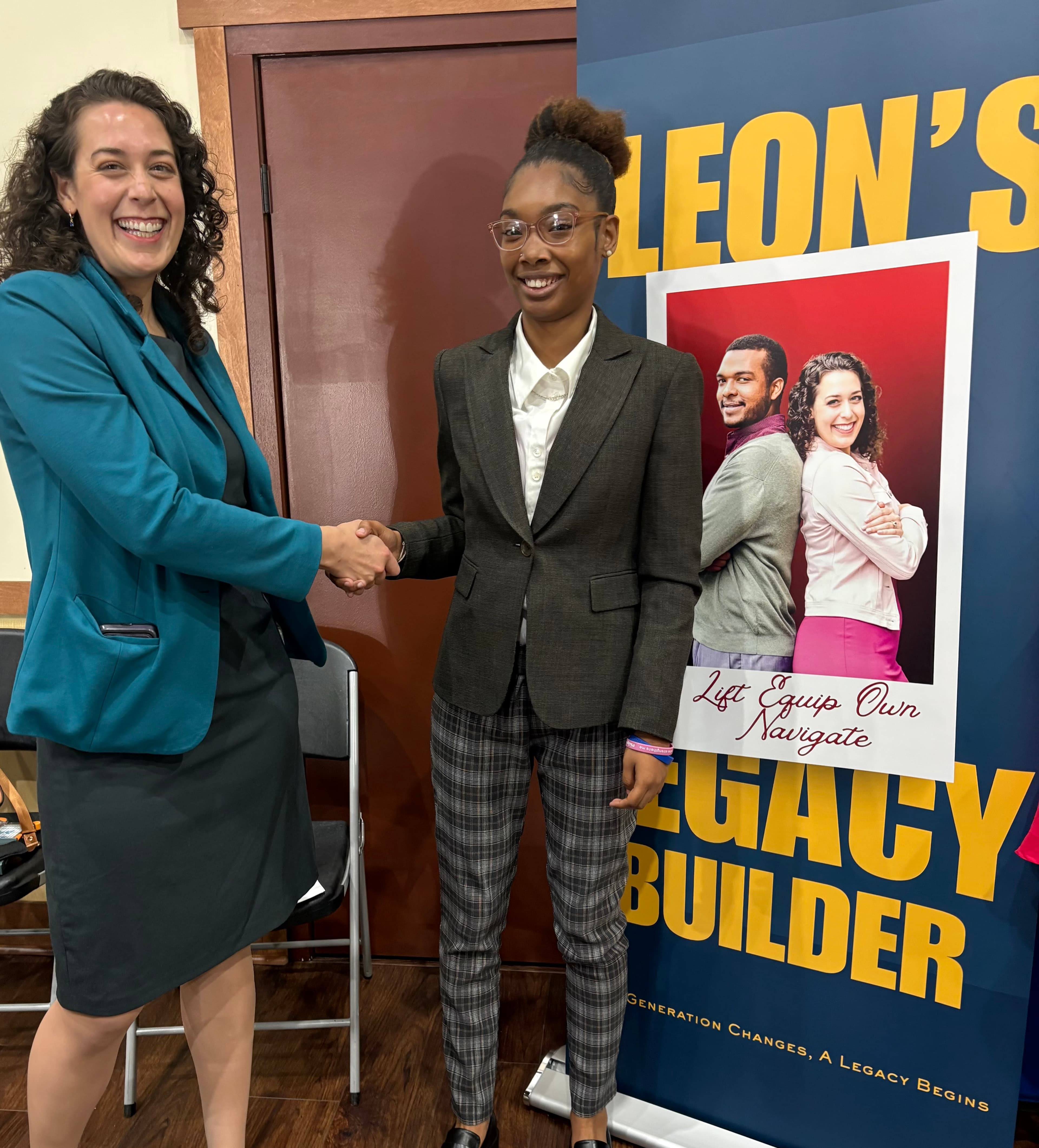 Two smiling women in professional attire shake hands in front of a promotional banner.