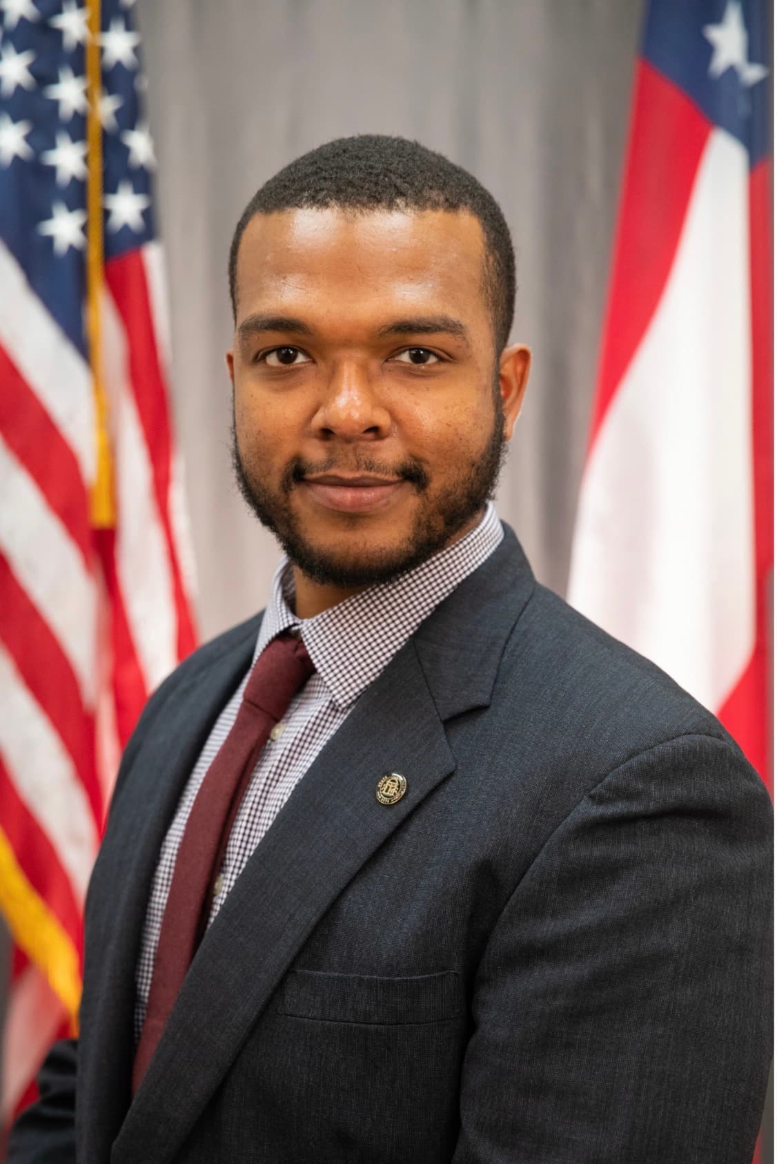 A Black man in a grey suit and maroon tie stands between two official flags.