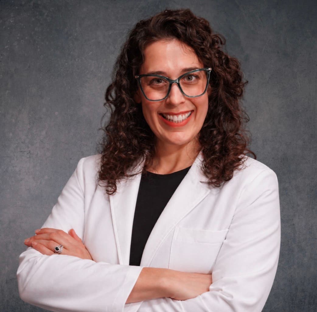 Smiling woman with curly brown hair and glasses wearing a white lab coat, arms crossed.