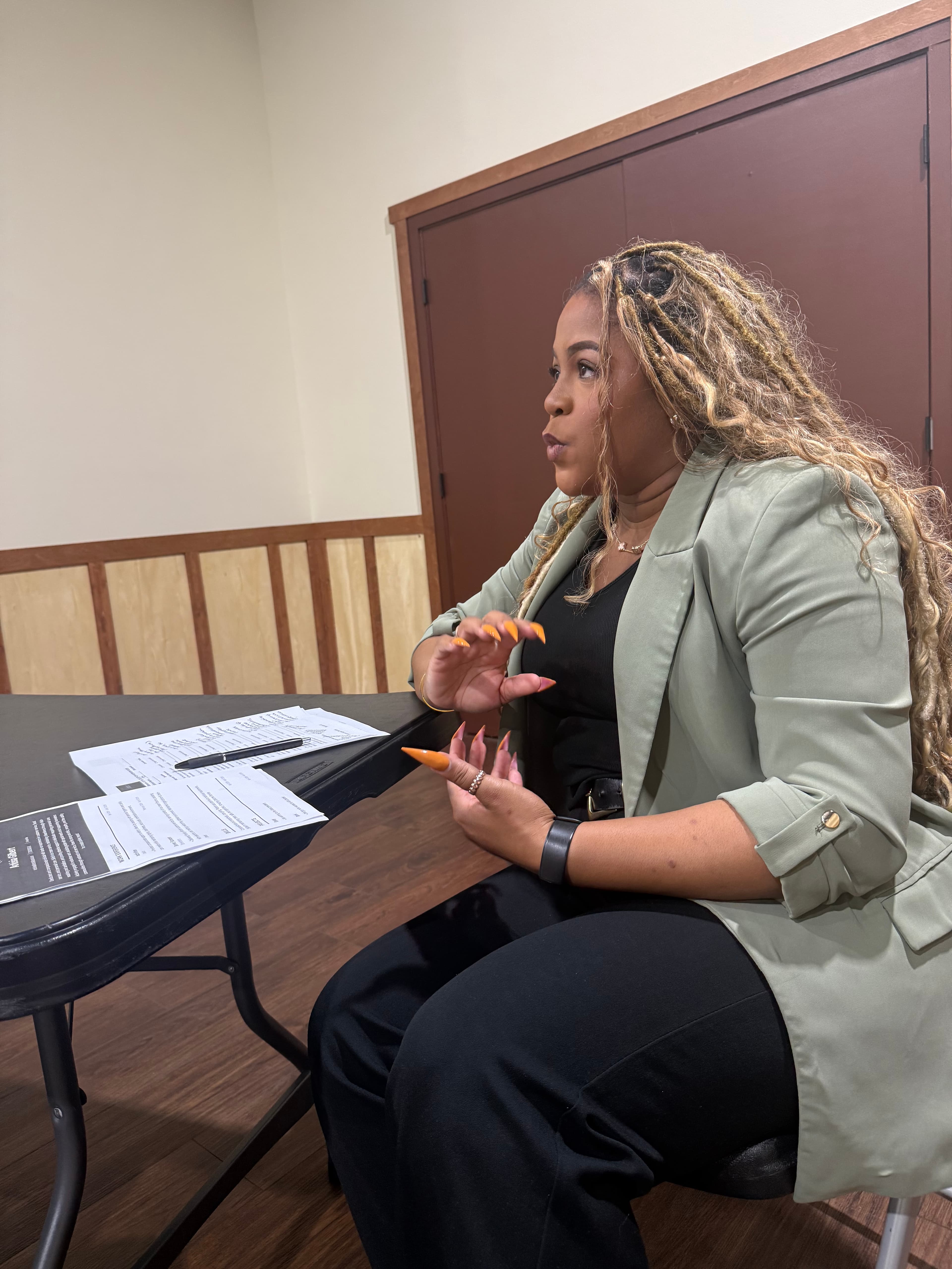 Woman with long braids and orange nails gestures while sitting at a table with documents.