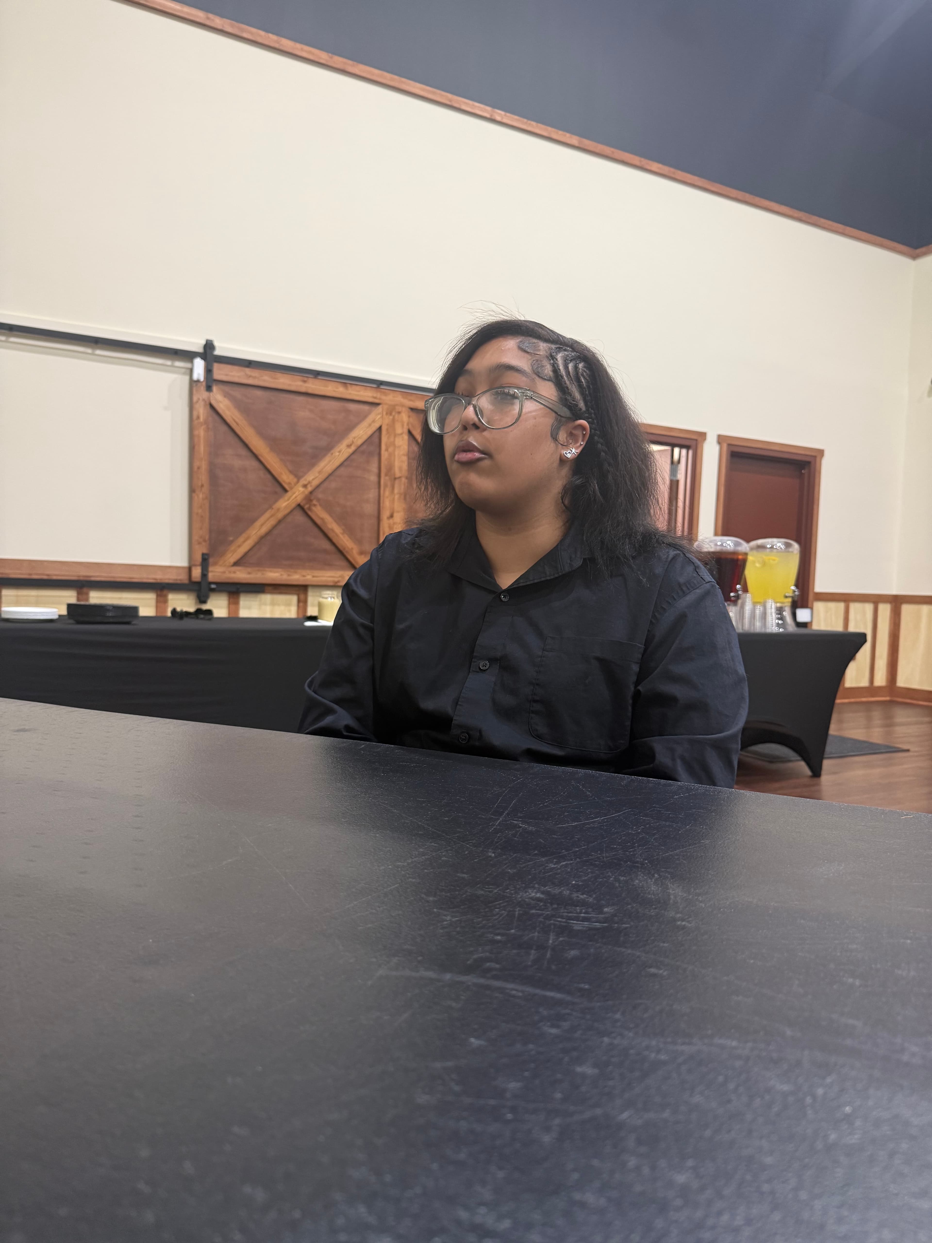 Woman wearing glasses and a black button-down shirt sits behind a black table in a hall.