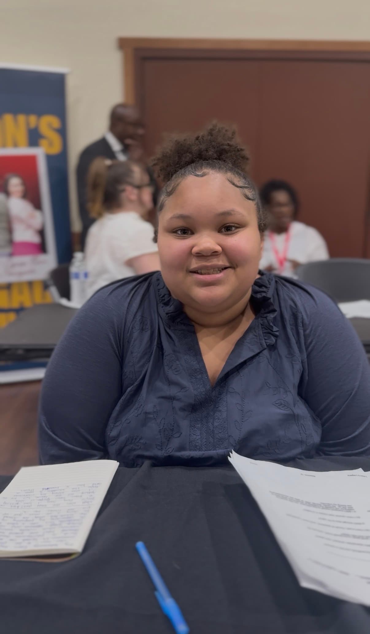 Smiling young woman sits at a table with a notebook, pen, and papers.