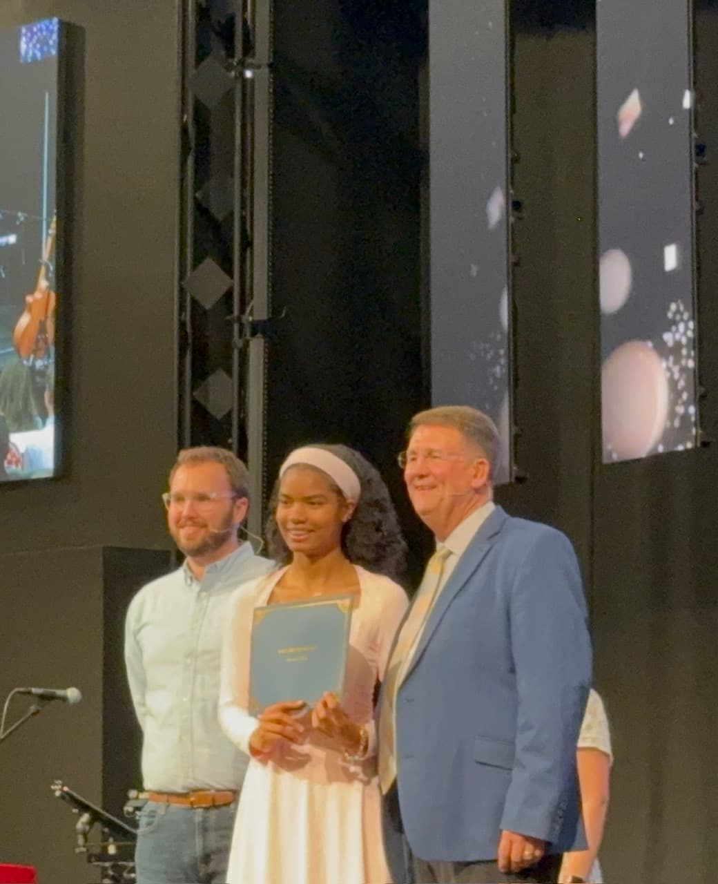 Smiling young woman holding a certificate folder, flanked by two men on a stage.