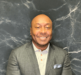Smiling bald Black man in a grey blazer and white shirt against a dark marble background.