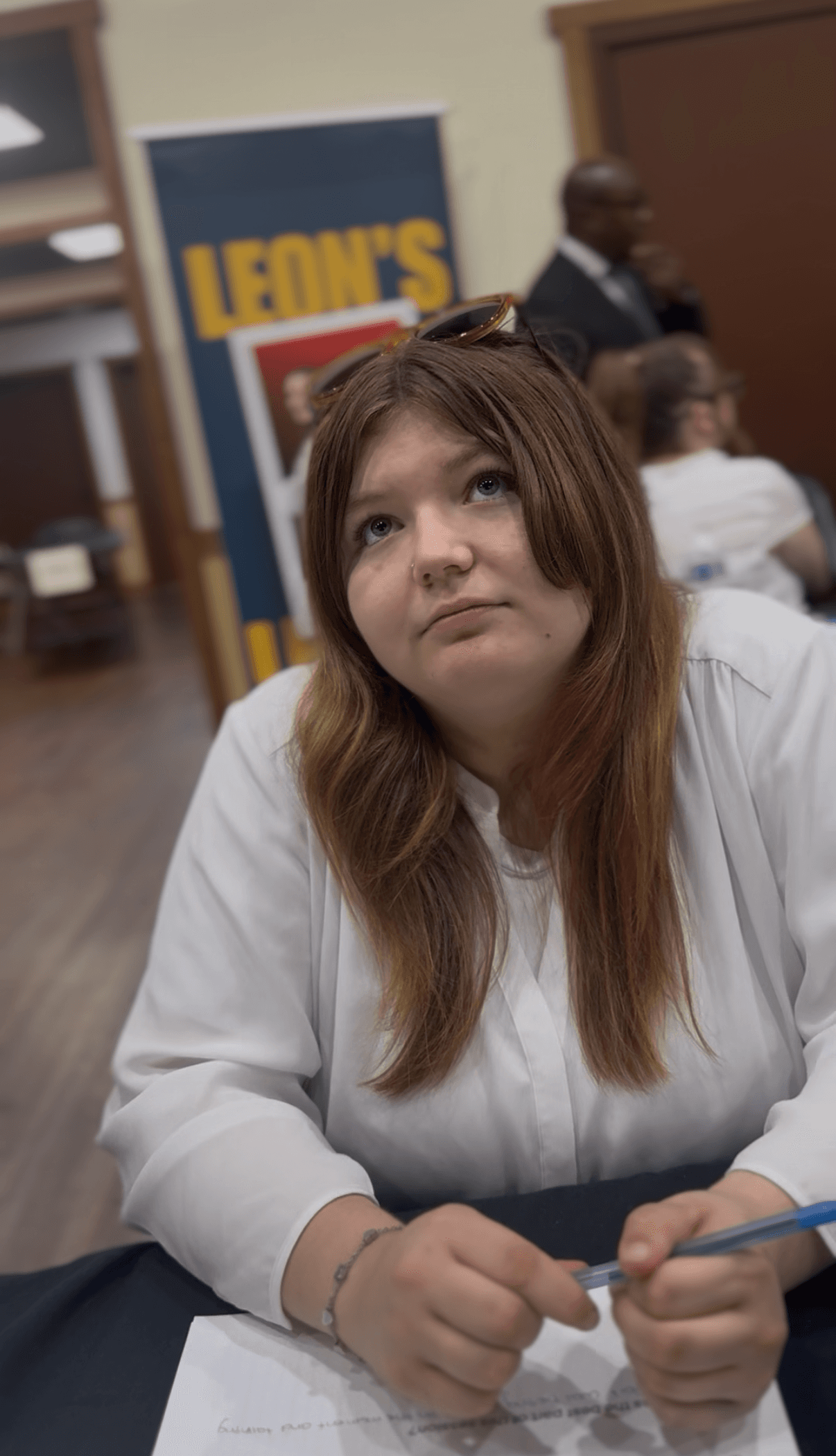Young woman with brown hair looks up thoughtfully while holding a pen over a paper.